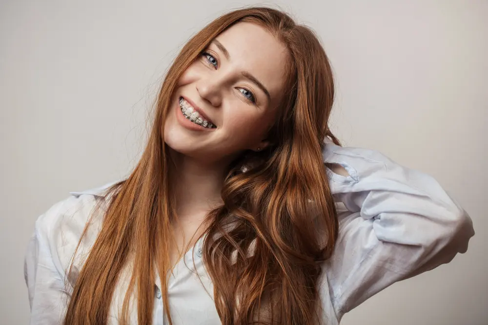 Smiling young woman with long red hair wearing clear braces and a white shirt at Wink Orthodontics, an orthodontist in Prosper and Colleyville, TX
