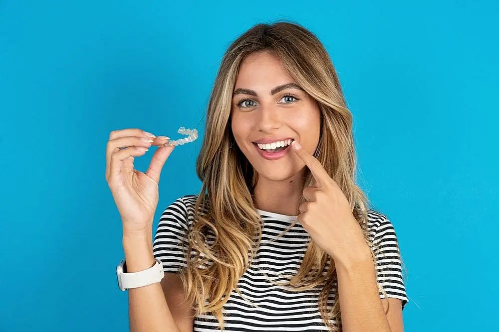 a woman against blue background pointing at her teeth and holding Invisalign clear aligners from Wink Orthodontics, an orthodontist in Prosper, TX 