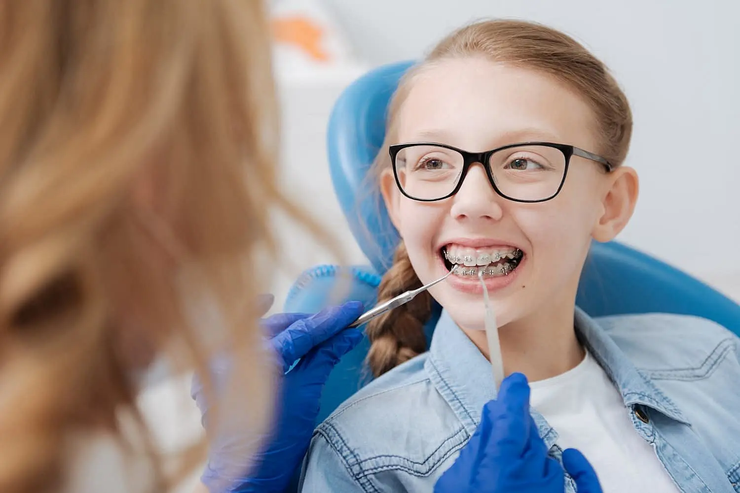A young girl with affordable braces smiles in a dental chair at Wink Orthodontics in Prosper or Colleyville TX, with dental equipment and a bright overhead light in the background.