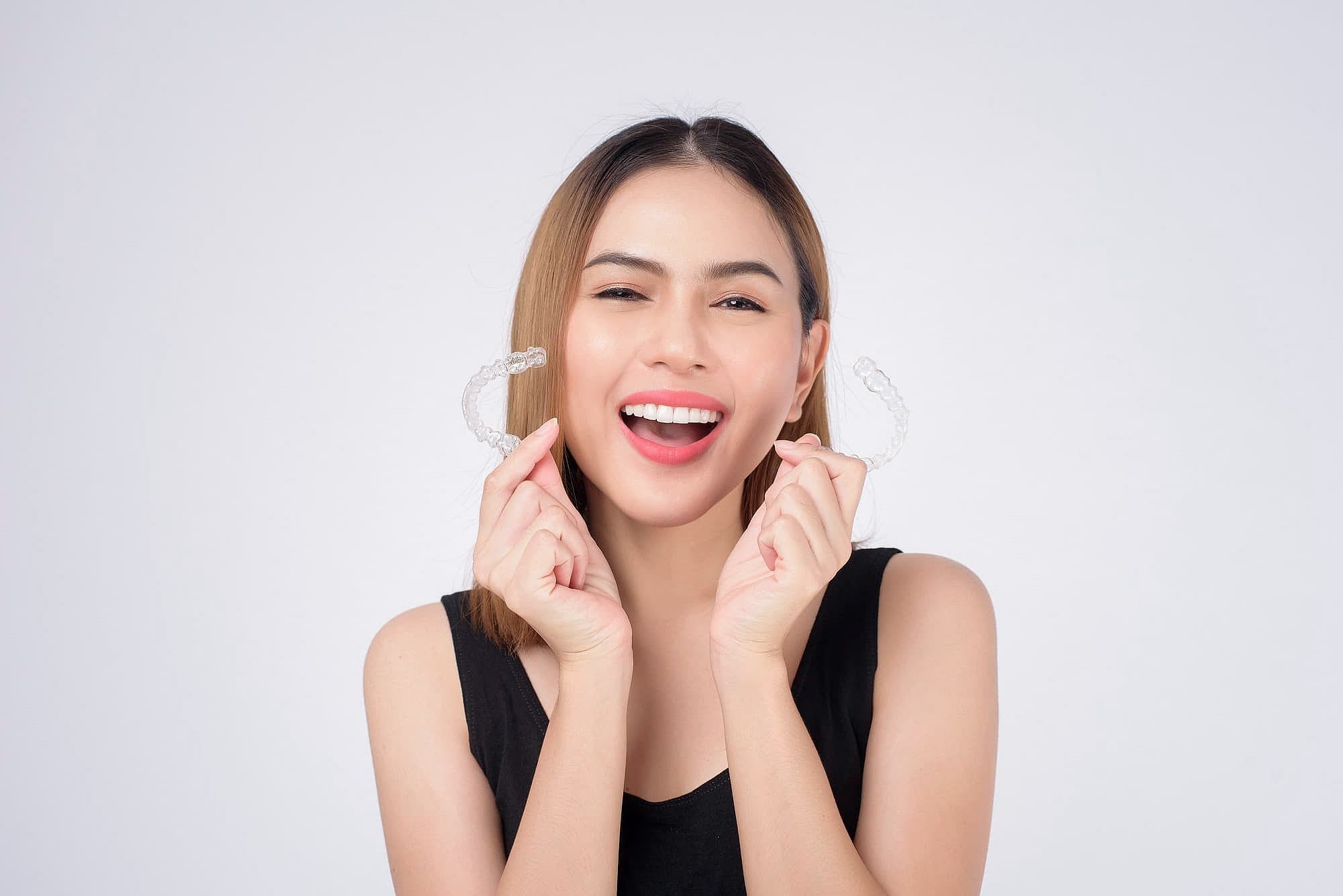 A woman smiles while holding a transparent dental aligner between their fingers, proud of their affordable Invisalign choice in Prosper or Colleyville, TX.
