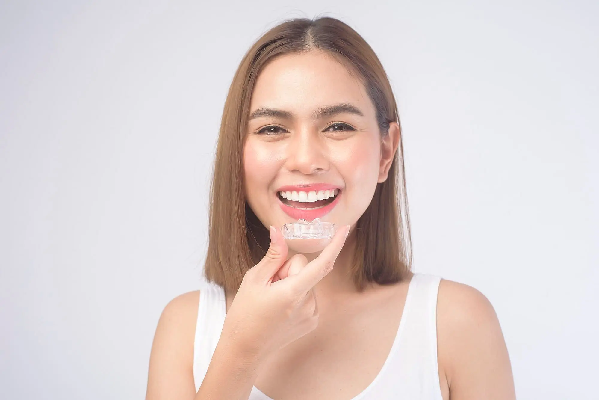 A woman in a white tank top smiles while holding Invisalign® Clear Aligners near her mouth against a plain white background at Wink Orthodontics in Colleyville, TX.