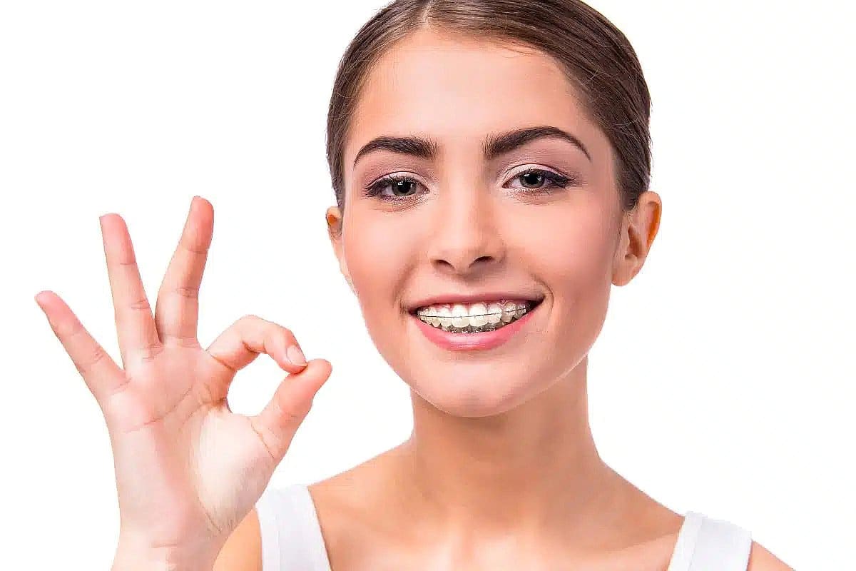A smiling young woman shows an OK hand gesture while wearing clear ceramic braces, representing Wink Orthodontics in Prosper, TX.