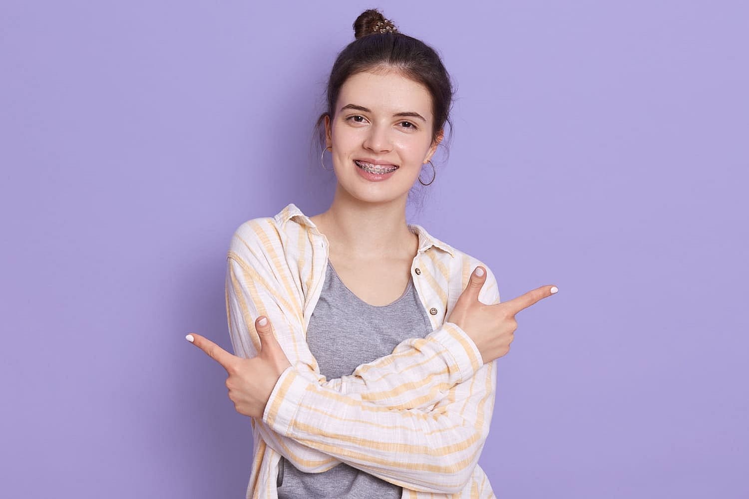 Smiling woman with braces for underbite treatment, points both index fingers to the sides on a purple background for Wink Orthodontics in Prosper or Colleyville, TX.