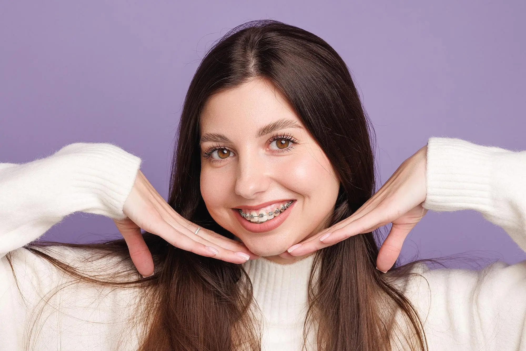 A young woman with long brown hair and braces smiles brightly, posing in a white sweater against a purple background at Wink Orthodontics in Colleyville, TX.