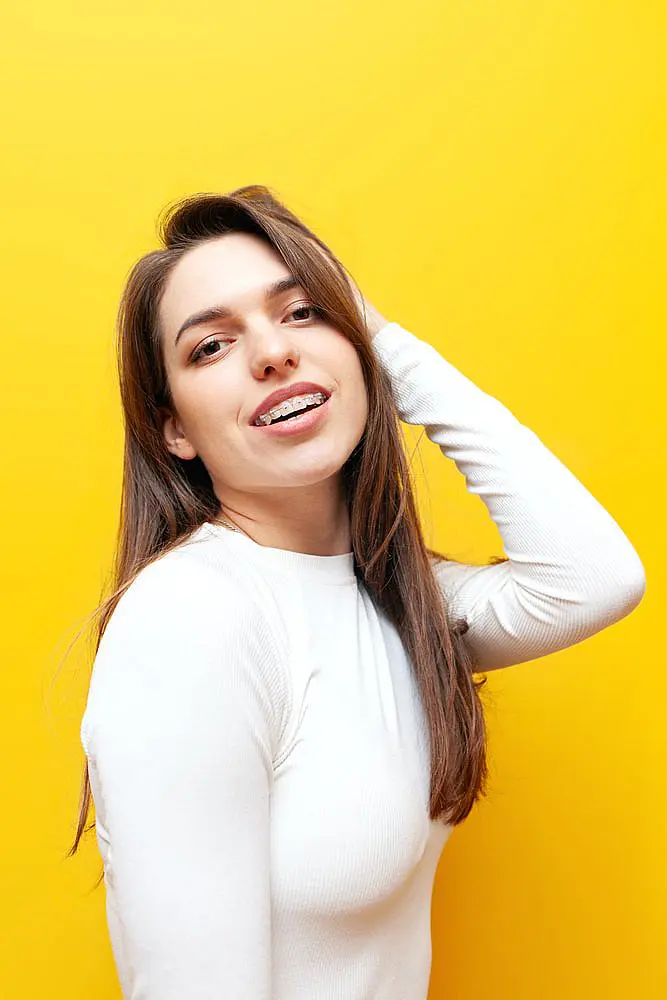 A young woman with long brown hair, wearing clear ceramic braces and a white long-sleeve top, smiles against a bright yellow background at Wink Orthodontics in Colleyville, TX.