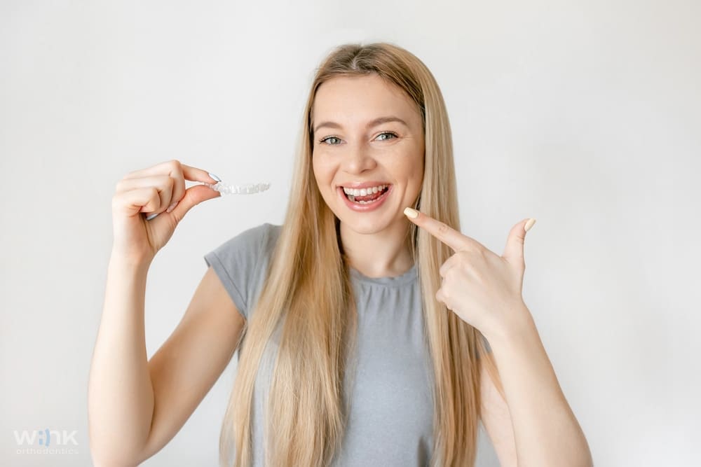 Happy woman with long blonde hair pointing to a clear aligner, demonstrating Invisalign use for straightening teeth while illustrating Does Insurance Cover Invisalign in Prosper and Colleyville, TX.