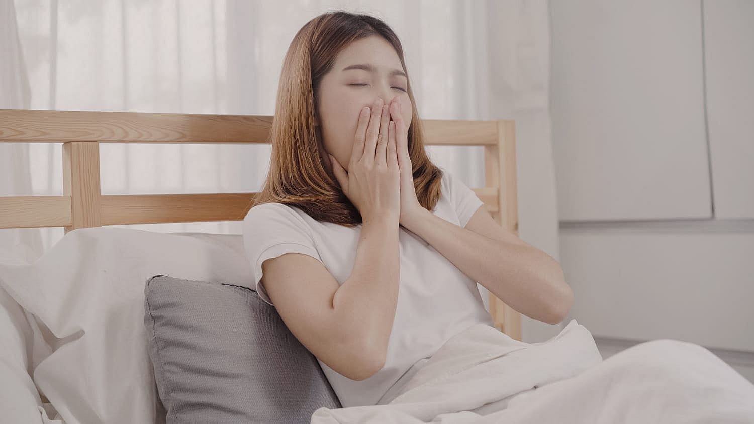 A woman sits in bed, a slight yawn escaping as she covers her mouth with both hands. She's clad in a white t-shirt, resting next to a gray pillow, looking very much the tired mouth breather in Prosper or Colleyville, TX