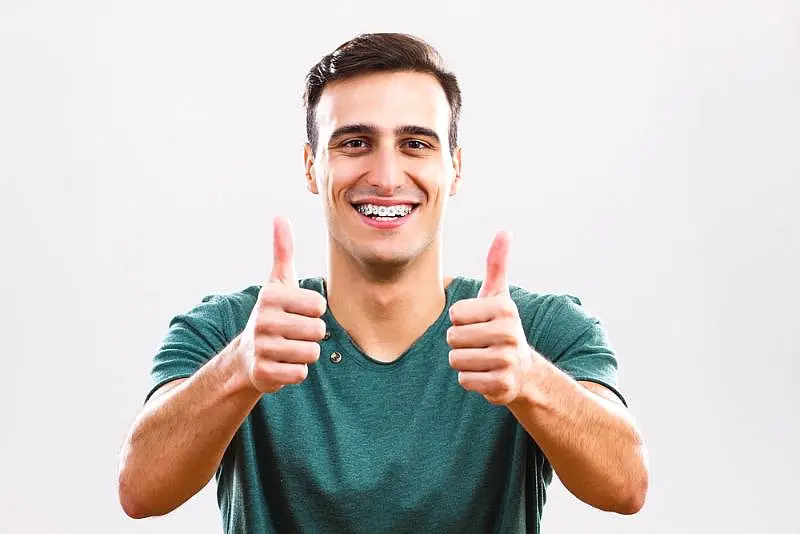 A young man with clear braces wearing a green shirt gives two thumbs up and smiles in front of a plain light background at Wink Orthodontics in Colleyville, TX.