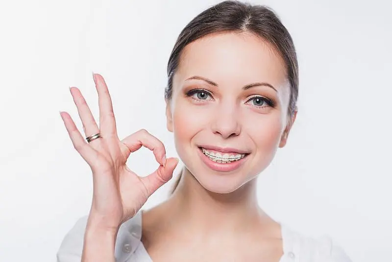 A woman with clear braces from Wink Orthodontics in Prosper, TX smiles and makes an "OK" hand gesture against a plain white background.