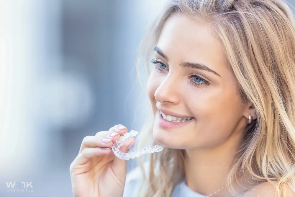 Smiling young woman holding a clear Invisalign aligner near her teeth, showcasing orthodontic treatment. while illustrating Does Insurance Cover Invisalign in Prosper and Colleyville, TX.