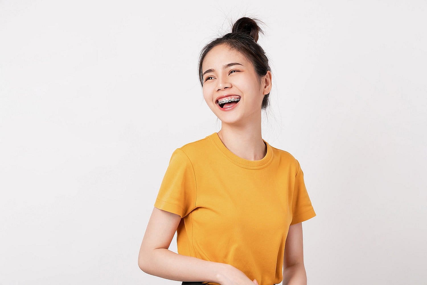 A young woman with braces, dressed in a yellow t-shirt, smiles confidently and glances to the side against a white background—showcasing results you can achieve at Wink Orthodontics in Colleyville, TX.