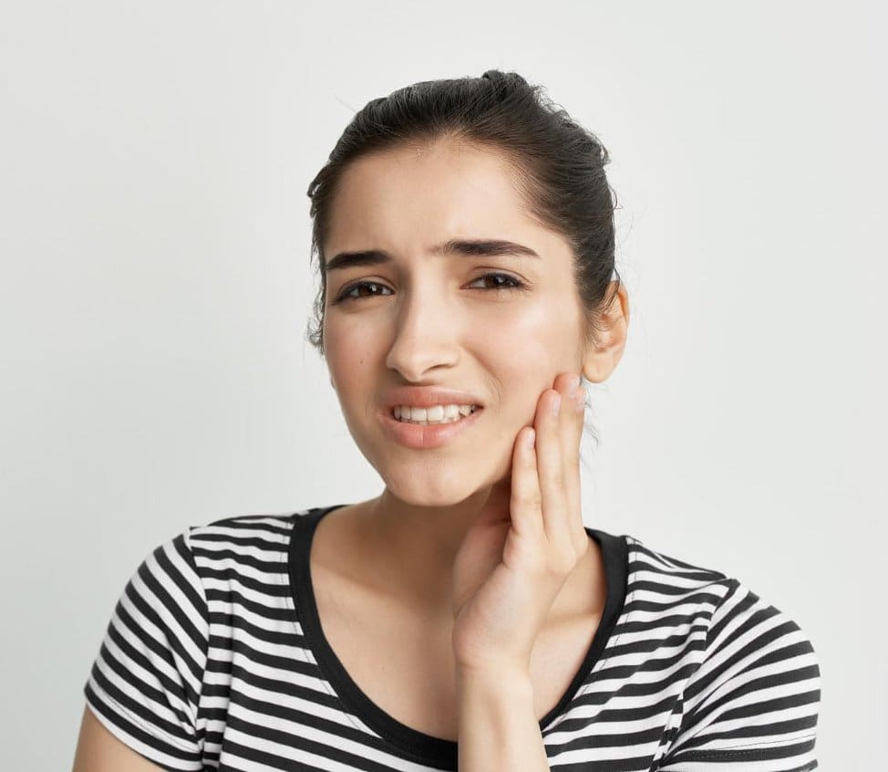 A young woman touches her cheek in discomfort, possibly from Overbite; Wink Orthodontics in Prosper or Colleyville, TX can help for Overbite treatment.