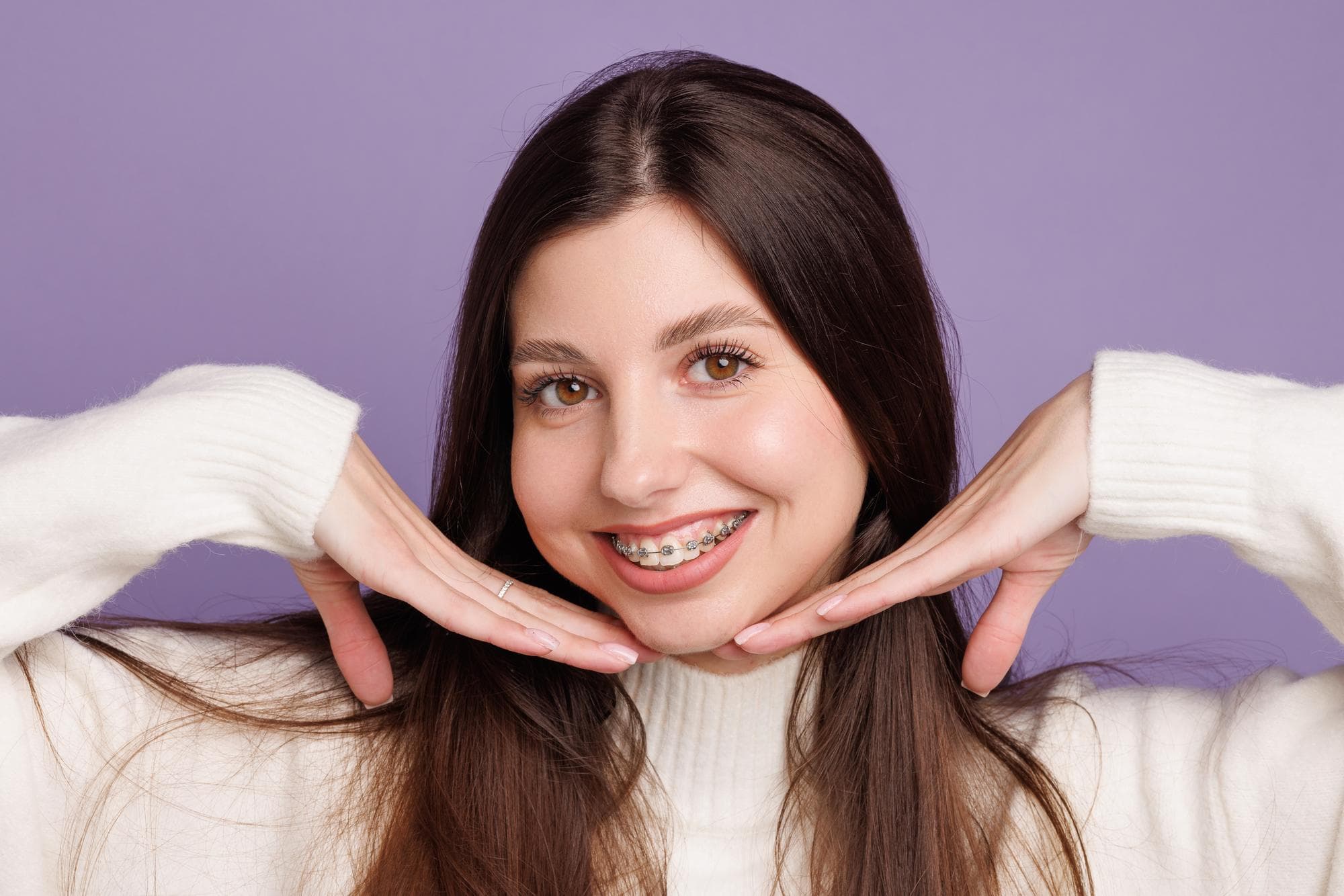 Smiling young woman with metal braces at Wink Orthodontics in Prosper or Colleyville, TX, poses in white sweater on purple background.