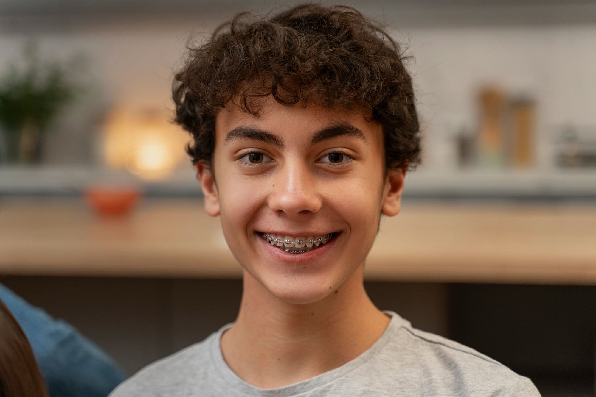 Smiling teen boy with curly brown hair and KLOwen Braces at Wink Orthodontics in Prosper TX, in a gray shirt.
