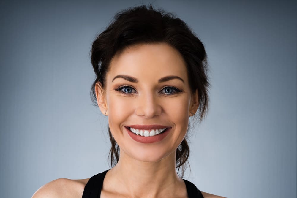 A confident woman with dark hair smiles at the camera after getting Ceramic Braces at Wink Orthodontics in Prosper, TX.