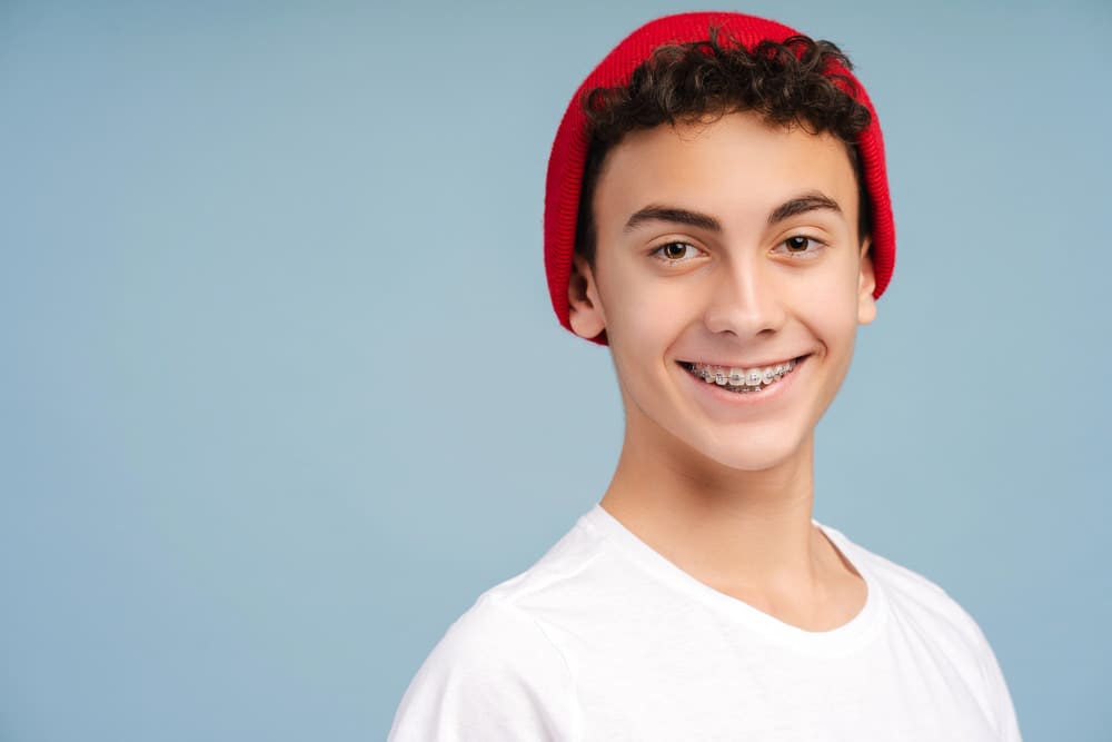 Wink Orthodontics Curly-haired teen in red beanie and white shirt smiles with braces, thinking about Braces Cost at Wink Orthodontics in Prosper or Colleyville, TX, on a light blue background.
