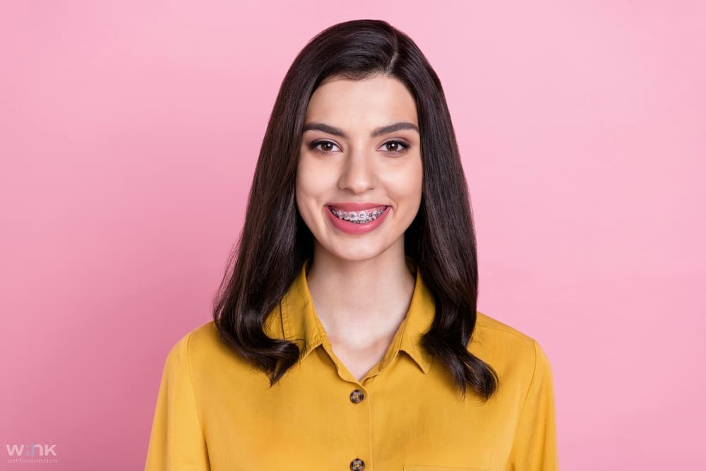 photo of nice optimistic brunette hair young lady wear metal braces pink background - How to Get Braces Glue Off Teeth in Prosper and Colleyville, TX