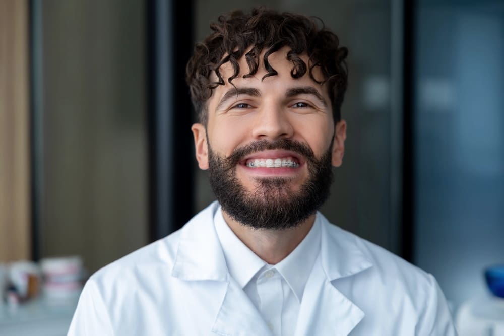 bearded-man-dentist-clear-braces-showing-importance-dental-care Smiling person with curly hair and a beard wearing a white lab coat, standing indoors - Clear Braces vs Metal Braces in Prosper and Colleyville, TX