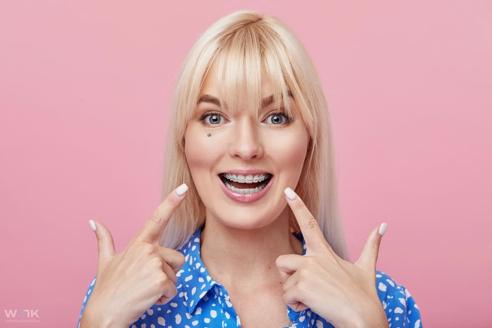 Blonde person smiling widely and pointing at their cheeks against a pink background - Clear Braces vs Metal Braces in Prosper and Colleyville, TX