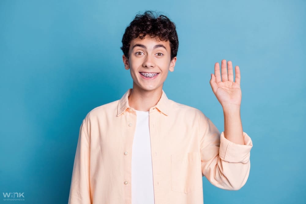 Person with short dark hair smiling and waving with one hand against a blue background - Clear Braces vs Metal Braces in Prosper and Colleyville, TX
