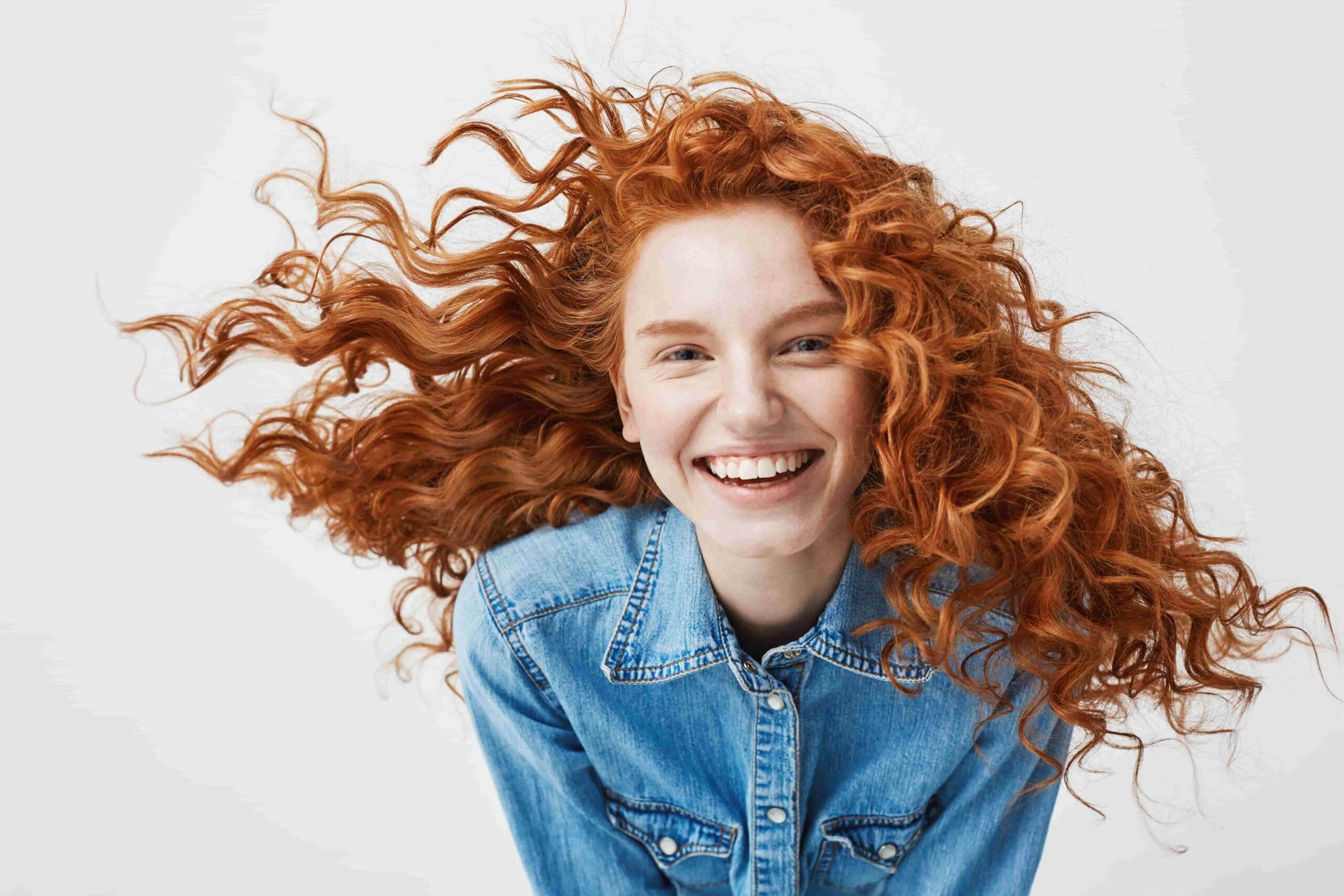 A smiling person with long, curly red hair and Brava Braces wears a blue denim shirt. Wink Orthodontics in Prosper or Colleyville, TX.