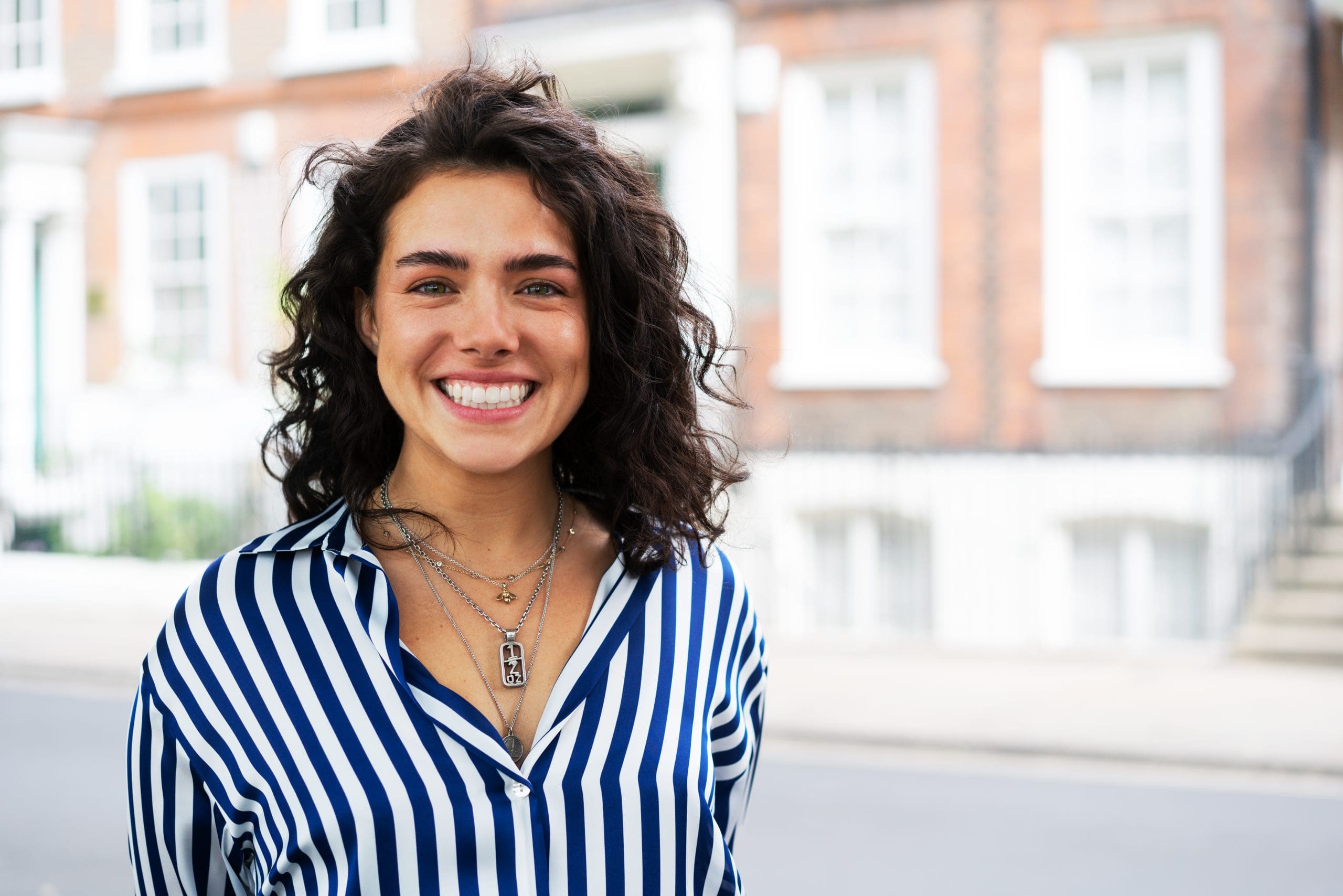 Smiling in front of brick buildings, a person with curly dark hair and Brava Braces from Wink Orthodontics in Prosper or Colleyville, TX wears a blue and white striped shirt and layered necklaces.